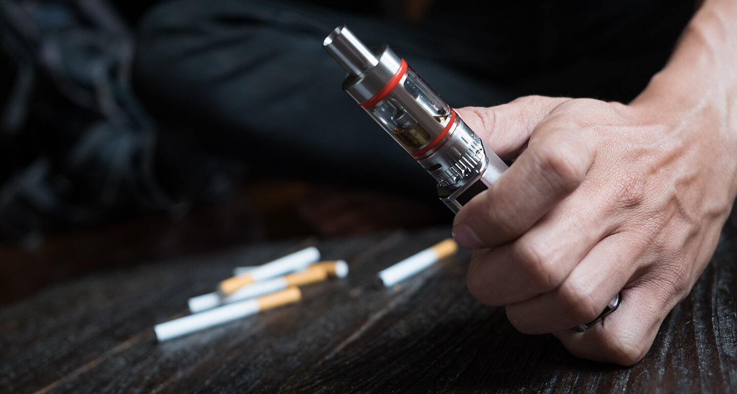 Eine Hand hält eine E-Zigarette während im Hintergrund Zigaretten auf dem Tisch liegen