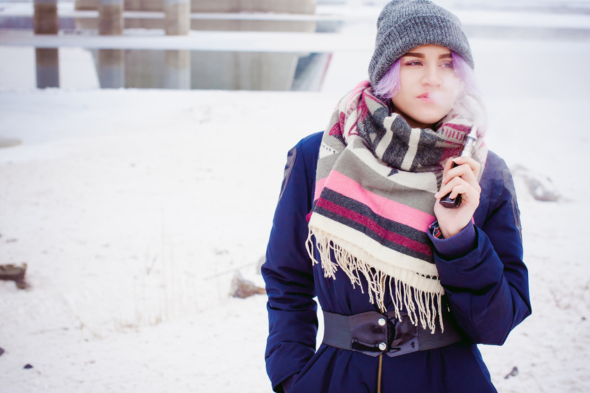 Frau mit grauer Mütze und Schal steht im Schnee und vapet, während sie eine E-Zigarette in der Hand hält