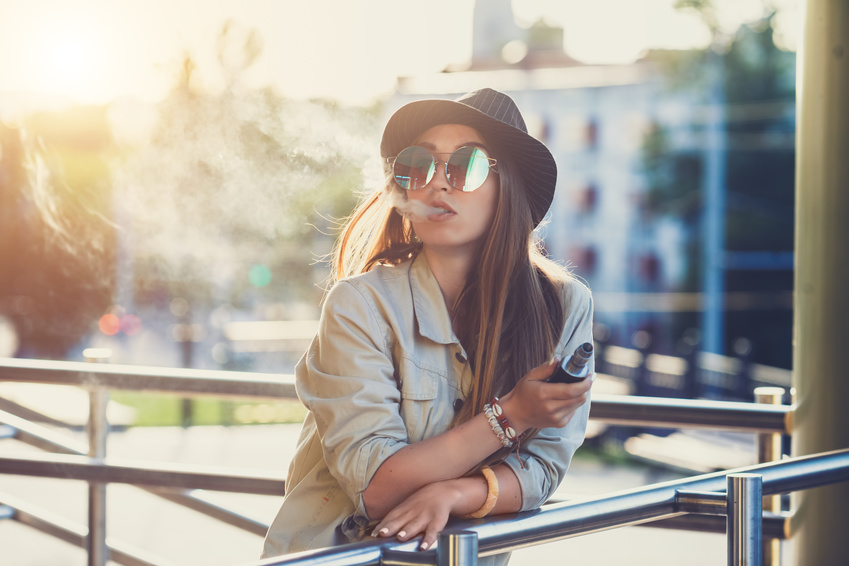 Junge Frau mit Sonnenbrille und Hut vapet an ein Geländer gelehnt